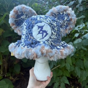 May include: A blue and tan crocheted bucket hat with two large fuzzy ears on top. The hat features a circular white patch on the front with a dark blue stylised symbol and a thin silver chain draped across the front and attached to the ears.