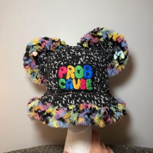 May include: A black and grey crocheted bucket hat with two large, fuzzy, multicoloured ears. The front features a patch with the embroidered text PROB CAUSE in bright colours. The hat is decorated with a small pink heart and a game controller charm.