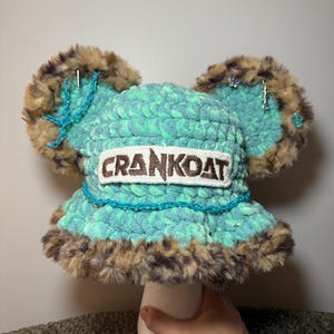 May include: A turquoise crochet bucket hat with two large, round, faux-fur ears in brown and black. The hat features a white rectangular patch with the text CRANKOAT in brown letters, a turquoise chain detail, and various small silver charms attached to the ears.