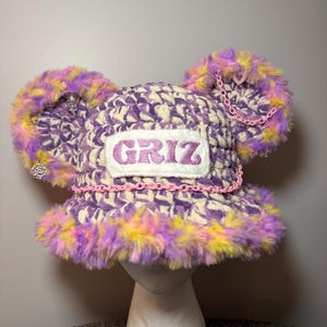 May include: A crochet bucket hat featuring a purple, yellow, and white variegated yarn pattern with large, fuzzy, multi-coloured ears. The hat includes a white rectangular patch with the text GRIZ, a pink plastic chain, and small charms.