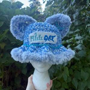 May include: A light blue and white crocheted bucket hat featuring plush faux fur ears, a fuzzy brim, and a rectangular patch on the front with the text ALL DAT in green and blue lettering. A thin teal chain is draped across the front of the hat.