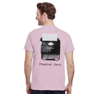 Op de afbeelding: Lichtroze t-shirt met een zwart-witte afbeelding van een vintage typemachine. De afbeelding bevat de tekst "Gazza" en "Chapter Zero". De typemachine heeft een blanco vel papier.