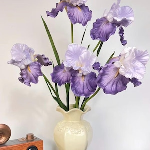 May include: A bouquet of five artificial iris flowers with ruffled petals in shades of light purple and white, featuring long green stems. The flowers are arranged in a cream-colored ceramic vase with a scalloped rim and embossed detailing.