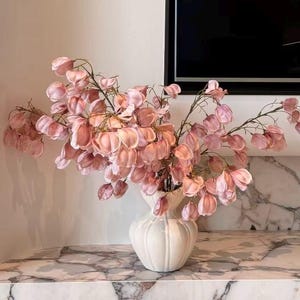 May include: A white ceramic vase with a ribbed, rounded body holds a large arrangement of delicate, pale pink, papery seed pods on thin, branching stems. The plant features numerous small, translucent, heart-shaped petals in soft blush tones.