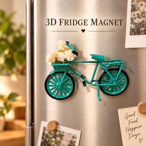 May include: A teal-coloured bicycle-shaped fridge magnet featuring a front basket filled with small, cream-coloured artificial flowers. The bicycle has black wheels, a teal frame, and a seat. The text 3D FRIDGE MAGNET is printed above the object.