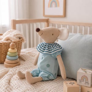 May include: A beige fabric mouse doll with a dark brown nose and large ears. The mouse wears a light blue romper featuring a darker blue polka dot pattern and a ruffled collar with blue and white horizontal stripes around the neck area.