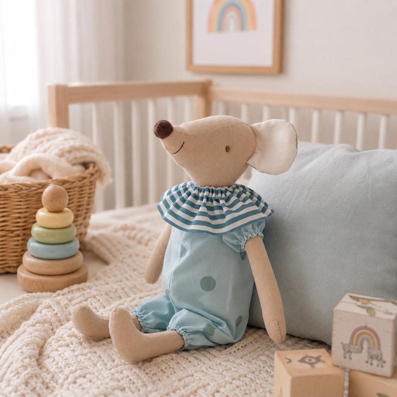 May include: A beige fabric mouse doll with a dark brown nose and large ears. The mouse wears a light blue romper featuring a darker blue polka dot pattern and a ruffled collar with blue and white horizontal stripes around the neck area.