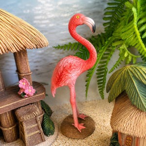 May include: A miniature pink flamingo figurine standing on a circular brown base. The flamingo has a long curved neck, a black-tipped beak, and textured feather details. It is positioned between a small wooden hut structure and green foliage.