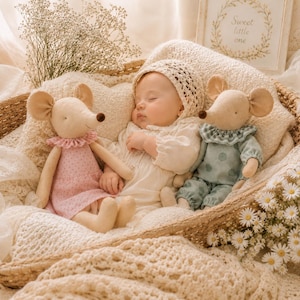 May include: A sleeping infant wearing a cream-colored bonnet and long-sleeved top, nestled between two plush mouse dolls. The mouse on the left wears a pink floral dress with a ruffled collar, while the mouse on the right wears a sage green polka-dot outfit.