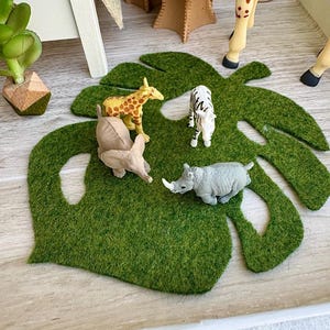 May include: A green felt monstera leaf mat featuring four small plastic animal figurines: a yellow and brown giraffe, a black and white zebra, a grey elephant, and a grey rhinoceros. The mat is shaped like a large tropical leaf with cutouts.