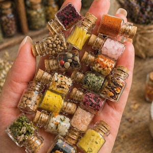 May include: A collection of small glass bottles, each containing different herbs and spices, held in a hand. The bottles have cork stoppers. The spices are in various colours, including red, yellow, green, and brown. The arrangement is visually appealing.