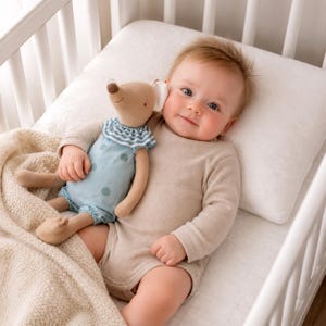 May include: A plush mouse toy with a tan body, wearing a light blue romper featuring dark blue polka dots and a blue and white striped ruffled collar, held by a baby wearing a beige long-sleeved ribbed bodysuit while lying on a white pillow.