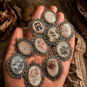 Könnte beinhalten: Eine Sammlung von neun ovalen Anhängern mit verzierten bronzefarbenen Metallrahmen und filigranen Kanten. Jeder Anhänger zeigt einen einzigartigen bedruckten Einsatz, darunter florale Muster, eine alte Handschrift und verschiedene Illustrationen.