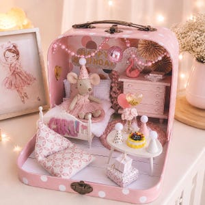 May include: A miniature dollhouse inside a pink polka-dot suitcase featuring a plush mouse in a party hat sitting on a bed. The room includes a white table with a small cake, gift boxes, a dresser, a flamingo figurine, and a banner reading HAPPY BIRTHDAY.