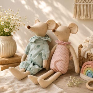 May include: Two beige fabric mouse dolls with long limbs and embroidered facial features. One mouse wears a light blue romper with large green polka dots and a ruffled collar, while the other wears a pink romper with small dots and a ruffled collar.