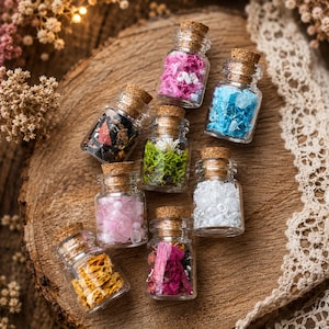 May include: Eight small clear glass bottles with cork stoppers, each filled with different materials including pink, blue, and white crystals, green moss, dried flower petals, and dark botanical pieces, arranged in a cluster on a wooden surface.