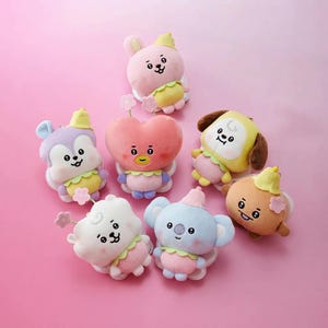 May include: A collection of seven small plush toys featuring various cartoon characters with pastel colors, including a pink heart, a blue koala, a white cloud, a yellow dog, a brown creature, a purple animal, and a pink rabbit, all with flower accents.