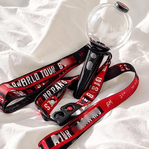 May include: A black and clear light stick with a clear globe top and a red and black lanyard. The lanyard has white text that reads "WORLD TOUR" and other text. The light stick has two buttons.