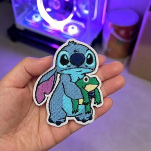 以下が含まれることがあります： 大きな耳と濃い青色の鼻を持つ水色のエイリアンのキャラクターが、黄色いお腹をした小さな緑色のカエルを抱えている刺繍ワッペン。ワッペンには白い縁取りがあり、2つのキャラクターの輪郭に沿った形状をしています。