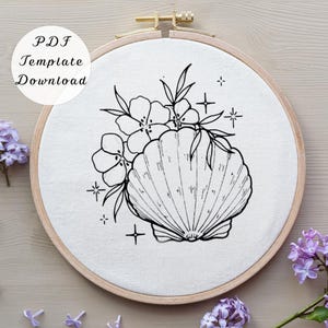 May include: A black and white line drawing of a seashell and flowers is centered on a white fabric stretched within a wooden embroidery hoop. The text "PDF Template Download" is in a white circle in the upper left. The design includes floral and starburst elements.