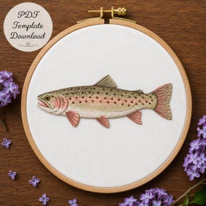 May include: A circular wooden embroidery hoop containing white fabric with a detailed thread-stitched rainbow trout. The fish features a tan and olive body with small dark spots, a pinkish lateral stripe, and pink-tinted fins. Text reads PDF Template Download.