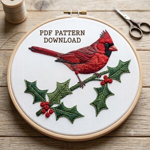 May include: A round wooden embroidery hoop containing white fabric with a hand-stitched red cardinal bird perched on a green holly branch with red berries. The text PDF PATTERN DOWNLOAD is printed in brown above the bird. A spool and scissors are nearby.