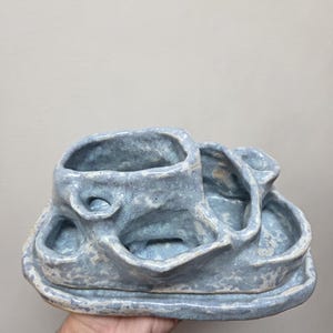 Op de afbeelding: Een lichtblauwe keramische organizer met een gespikkelde, getextureerde afwerking. Het object heeft meerdere onderling verbonden compartimenten van verschillende hoogtes en onregelmatige, organische vormen op een enkele, verhoogde basis.