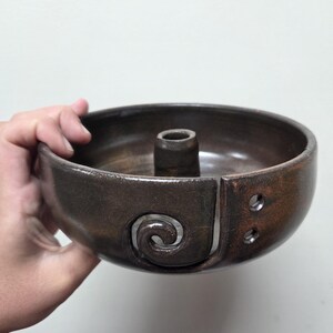 May include: A dark brown ceramic yarn bowl with a glossy finish. The bowl features a central post, a spiral-shaped cutout on the side for yarn feeding, and two small circular holes near the edge for organizing multiple strands of yarn.