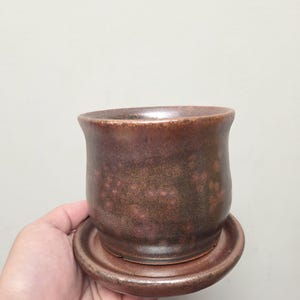 Könnte beinhalten: Ein zylindrischer Keramikübertopf mit ausgestelltem Rand und passendem runden Untersetzer. Die Keramik weist eine gesprenkelte, dunkelbraune und rötlich-bronzefarbene Glasur mit einem dezenten metallischen Glanz und einer glatten Oberfläche auf.