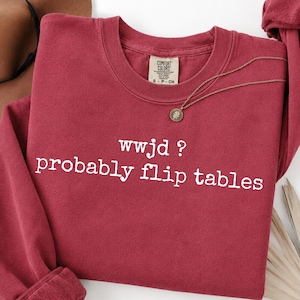 May include: A dark red crewneck sweatshirt featuring the white text "wwjd ? probably flip tables" printed on the front. The garment has a Comfort Colors brand label at the inner neckline and long sleeves with ribbed cuffs and a ribbed hem.