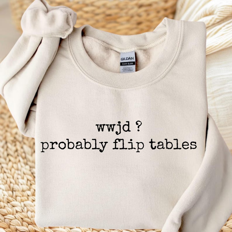 May include: A sand-colored crewneck sweatshirt featuring the black text "wwjd ? probably flip tables" printed on the front. The garment has long sleeves, a ribbed collar, and a Gildan brand label visible inside the neckline of the fleece-lined fabric.
