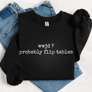 May include: A black long-sleeve sweatshirt featuring white text printed on the front that reads: wwjd ? probably flip tables. The garment has a crew neckline and ribbed cuffs, displayed against a pair of blue denim jeans.