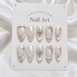 May include: A set of ten almond-shaped press-on nails with a pearlescent white finish, displayed on a card labeled COLLECTION Nail Art. The nails feature silver starburst charms, small white pearls, clear rhinestones, and intricate white lace patterns.