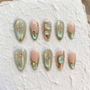May include: A set of ten artificial nails in various shapes and designs. The nails feature a combination of light green, pink, and gold accents. Some nails have starburst and gemstone details, while others have gold swirls.