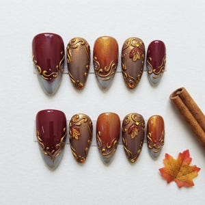 Può includere: Un set di dieci unghie finte con una sfumatura nei toni del bordeaux intenso, bronzo e arancione. Ogni unghia è decorata con filigrana dorata in rilievo, e alcune includono piccoli dettagli a forma di foglia e minuscoli strass incastonati.