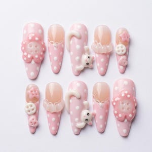 May include: A set of ten pink and white polka dot artificial nails. Each nail features unique embellishments, including 3D flowers, bunny figures, and lace details. The nails are almond-shaped and set against a white background.