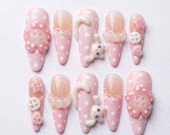 May include: A set of ten pink and white polka dot artificial nails. Each nail features unique embellishments, including 3D flowers, bunny figures, and lace details. The nails are almond-shaped and set against a white background.