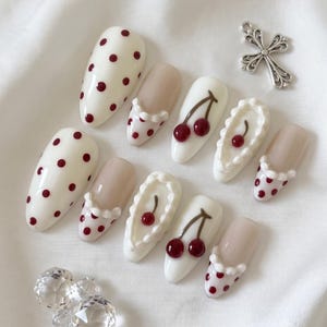 May include: A set of ten press-on nails featuring a mix of designs including cream-colored bases with dark red polka dots, nude bases with white ruffled accents, and white bases with painted red cherry motifs, accompanied by a silver cross charm.