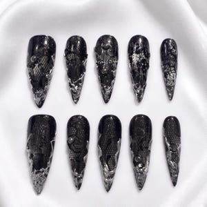 May include: A set of ten press-on nails featuring a glossy black base with intricate black lace patterns, accented by metallic silver liquid-like textures and small spherical studs, arranged in two rows of five on a white surface.
