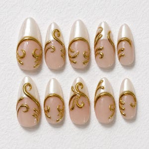 May include: A set of ten almond-shaped press-on nails featuring a light pink base with a pearlescent white tip. Each nail is decorated with raised, metallic gold filigree swirls that extend from the base toward the center of the nail.
