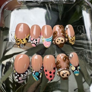 May include: A set of ten artificial nails in a clear plastic case. The nails feature a nude base with various designs, including a monkey, polka dots, and floral patterns. Some nails have rhinestones for added detail. The designs incorporate colors like brown, pink, yellow, and black.
