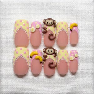May include: A set of ten press-on nails with a light pink base and a yellow polka dot design. Some nails feature a monkey or banana embellishment. The nails have a French tip design with a white lace trim.