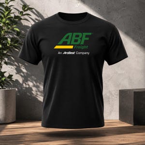 May include: A black short-sleeved t-shirt featuring the green and yellow ABF Freight logo on the chest, with the text "An ArcBest Company" printed in white directly below the logo. The shirt has a crew neckline and a standard fit.