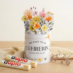 Könnte beinhalten: Ein zylindrischer Behälter mit gelben und orangen Blumenillustrationen und dem Text "FÜR EINE TOLLE LEHRERIN DANKE FÜR ALLES." Der Behälter enthält Papierblumen-Ausschnitte, begleitet von zwei weißen Duplo-Schokoriegeln.