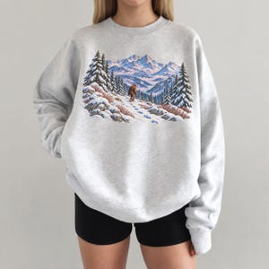 May include: A light gray crewneck sweatshirt featuring a graphic print of a brown creature walking through a snowy mountain landscape with evergreen trees, rocky terrain, and blue-tinted peaks under a soft, light-colored sky.