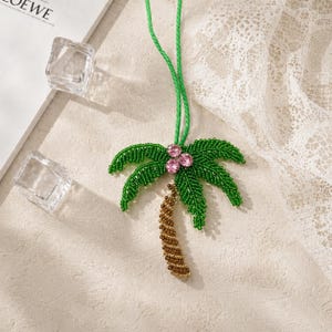 Op de afbeelding: Een kralen ketting met een palmboomhanger. De palmboom heeft groene, met kralen bezette bladeren, een bruine, met kralen bezette stam en roze edelsteenaccenten. De ketting heeft een groen koord. De hanger is een accessoire met een tropisch thema.