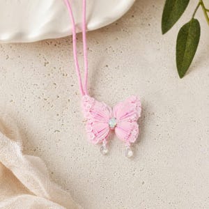 Puede incluir: Un collar con colgante de mariposa rosa. El colgante de mariposa tiene una gema iridiscente central y dos colgantes transparentes en forma de lágrima. El cordón del collar es rosa.