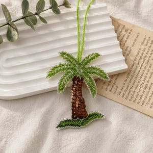 Peut inclure: Pendentif en forme de palmier perlé, avec un tronc marron à paillettes, des frondes vertes perlées et un cordon vert. Posé sur une surface blanche avec des feuilles vertes et un papier avec du texte.