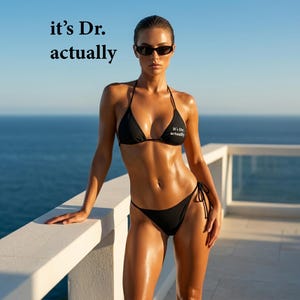 Op de afbeelding: Een vrouw in een zwarte bikini en zonnebril staat op een wit balkon met uitzicht op de oceaan. De bikinitop heeft de tekst "it's Dr. actually". De lucht is blauw en zonnig.