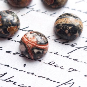 May include: Close-up of five round, polished, multi-colored stones with a black, white, red, and brown marbled pattern. The stones are on a white surface with black ink writing.